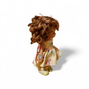 May include: A vintage doll with curly brown hair, wearing a yellow dress with a butterfly pattern and a pink ribbon. The doll has yellow shoes and is posed against a white background.