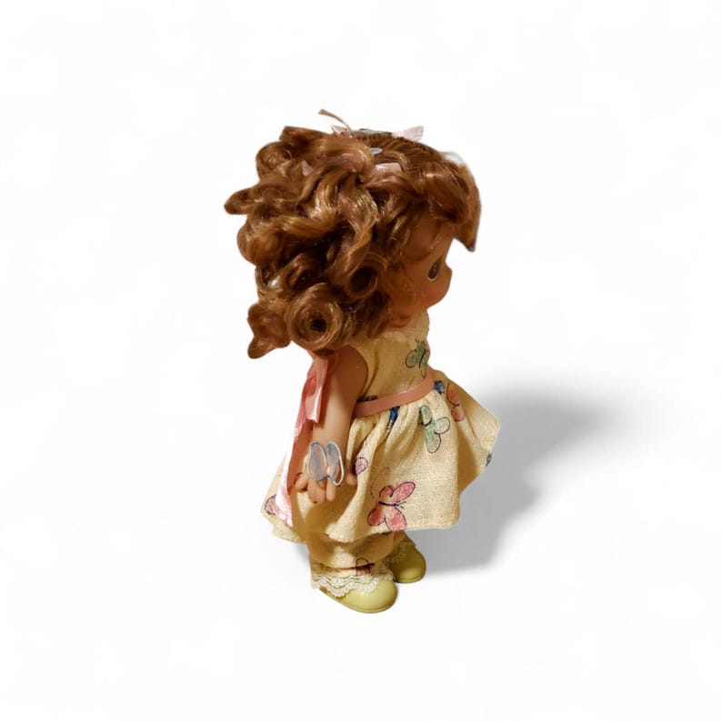 May include: A vintage doll with curly brown hair, wearing a yellow dress with a butterfly pattern and a pink ribbon. The doll has yellow shoes and is posed against a white background.