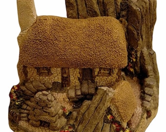 Może przedstawiać: Miniaturowy kamienny domek z strzechowym dachem osadzony jest wśród skalistych formacji. Domek ma komin i małe drzwi. Skały są pomalowane w odcieniach brązu i szarości, a wokół podstawy domku rozsiane są małe czerwone i żółte kwiaty.