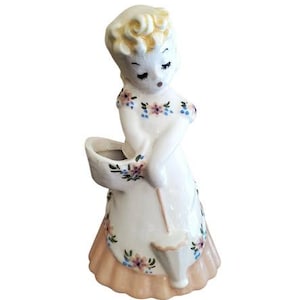 May include: A vintage ceramic figurine of a woman in a white dress with floral accents. The figurine has a peach-colored skirt and a basket. The woman's hair is a light yellow color.