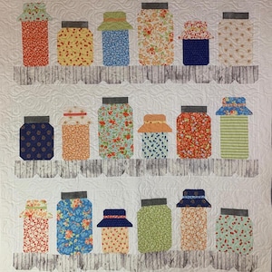 May include: A quilted wall hanging featuring a design of various jars with lids, arranged on wooden shelves. The jars are filled with colorful floral and patterned fabrics, including red, yellow, blue, and green. The background is white with quilted details.