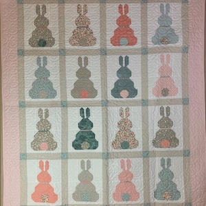 May include: A handmade quilt featuring a repeating pattern of bunny silhouettes in various colors and patterns. The quilt has a light pink border and a neutral-toned grid, with each bunny block set against a white background. The bunnies have floral, polka dot, and solid designs.