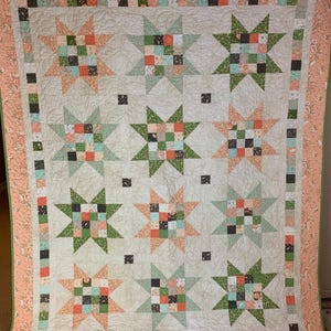 May include: A handmade quilt featuring a star pattern in shades of green, peach, and blue on a cream background. The quilt has a floral border in peach and green. The star blocks are made of small squares of fabric.
