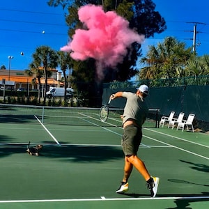 May include: A man in a white hat and green shorts plays tennis on a green court. He hits a pink powder-filled ball that explodes into a cloud of pink smoke. A small dog watches from the sidelines.