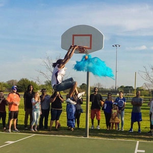 May include: A group of people gathered around a basketball hoop, watching a person dunk a basketball. Blue powder explodes from the ball as it goes through the hoop.