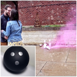 May include: A black bowling ball with three silver dots on the surface. The ball is sitting on a white surface. In the background, a couple is standing in front of a brick wall. There are five white bowling pins on the ground, and a pink cloud of smoke is behind them.