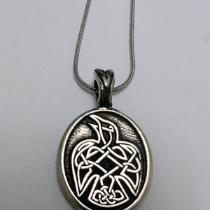 Puede incluir: Collar con colgante plateado con un diseño de nudo celta en forma de ave. El colgante ovalado cuelga de una fina cadena plateada. El diseño del ave es intrincado, con un acabado texturizado. El collar se muestra sobre un fondo blanco.