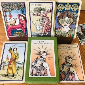 May include: A set of tarot cards with colorful illustrations and gold accents. The cards are from the "Tarot of the Sevenfold Mystery" deck, second edition by Robert M. Place. The cards feature images of a queen, a blindfolded woman, a man with a staff, and a woman with stars around her eyes.