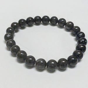 May include: A dark gray beaded bracelet. The bracelet is made of round, polished beads with subtle variations in color and texture. The beads are strung together on an elastic band, creating a flexible and comfortable fit. The bracelet is a simple yet elegant accessory.
