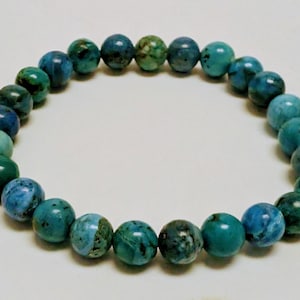 Op de afbeelding: Een armband met ronde, gepolijste kralen in tinten van teal, turquoise en diepgroen. De kralen zijn aan elkaar geregen en vormen een cirkelvorm. De kralen hebben een gemarmerd uiterlijk. Sieraad.