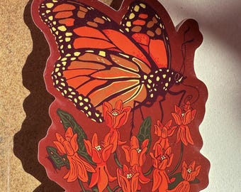 May include: A sticker featuring a monarch butterfly with orange and black wings, perched on a cluster of orange flowers and green leaves. The sticker has a red background and a detailed illustration style.