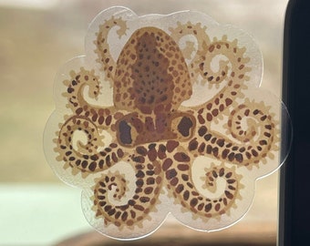 May include: A transparent sticker featuring a detailed brown octopus design. The octopus has a textured body and curling tentacles, with a decorative border. The sticker is affixed to a window, with a blurred outdoor scene visible in the background.