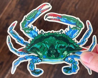 May include: A sticker featuring a detailed illustration of a blue crab. The crab has a green body with blue and red accents on its claws and legs. The sticker is held against a dark wood grain background.