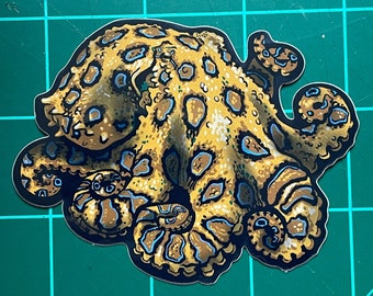 May include: A sticker featuring a detailed illustration of a blue-ringed octopus. The octopus has a yellow-brown body with numerous dark brown and blue spots. The tentacles are curled, and the sticker has a white border.