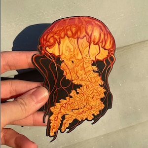 May include: A vibrant sticker featuring a detailed illustration of a jellyfish. The jellyfish has a reddish-orange bell and flowing tentacles in shades of orange and yellow, set against a black outline. The design is artistic and eye-catching.
