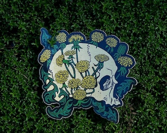 May include: A sticker featuring a skull design with yellow dandelions and green leaves. The skull is white with blue accents, and the dandelions are in full bloom. The sticker is set against a background of green foliage.