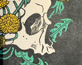 May include: Close-up of a graphic design featuring a skull with yellow dandelions and green leaves against a dark gray background. The skull is off-white with black eye sockets and nose. The dandelions are in full bloom.
