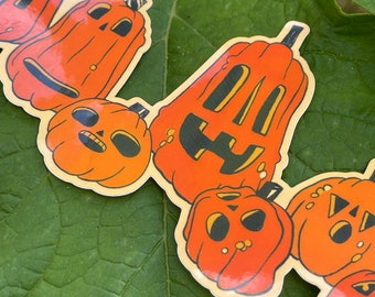 May include: A collection of orange pumpkin stickers with black facial features and stems. The stickers are in various shapes and sizes, with a vintage, cartoon-like design. The stickers are placed on a green leaf.