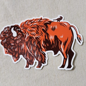 May include: A sticker featuring a stylized illustration of a bison in shades of orange and brown, with a small black bird perched on its back. The bison is depicted in profile, with detailed fur and horns.