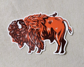 May include: A sticker featuring a stylized illustration of a bison in shades of orange and brown, with a small black bird perched on its back. The bison is depicted in profile, with detailed fur and horns.