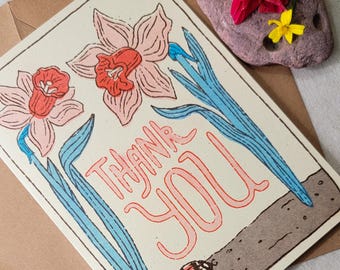May include: A cream-colored greeting card with a floral design and the words "THANK YOU" in coral. The card features pink daffodils with blue stems and a ladybug. The card is placed on a brown envelope.