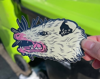 May include: A die-cut sticker featuring a stylized opossum with an open mouth, showing its teeth and pink tongue. The opossum has a cream-colored face and body with black and navy blue accents. The sticker is held against a blurred background.