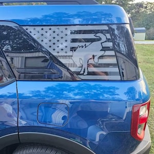 May include: A blue Ford Explorer with a black and white American flag decal on the rear window. The decal features a white silhouette of a bucking bronco.