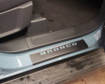 May include: A black and silver Bronco door sill plate installed in a vehicle. The word "BRONCO" is displayed in silver lettering on the black plate. The vehicle's interior includes a black seat and door panel.