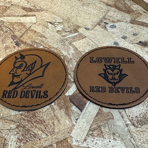 May include: Two brown leather patches with black stitching. Both patches feature a black and white illustration of a devil with horns and a red devil's tail. The text "Lowell Red Devils" is printed on both patches.