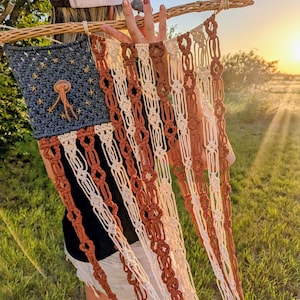 May include: A macrame wall hanging designed as a flag, featuring a dark blue square with embroidered gold stars and a central leather accent. The flag has alternating vertical stripes of cream and rust-colored knotted cord suspended from a wooden branch.