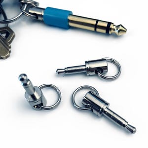 May include: A collection of silver-colored audio jack keychains. One keychain has a blue accent. The keychains are on a white background. The keychains are designed to look like audio jacks.