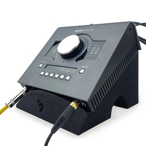 May include: A dark gray Apollo Twin X audio interface resting on a black angled stand. The device features a large central silver knob, multiple buttons, and digital level indicators. A yellow cable and a black cable are plugged into the front ports.