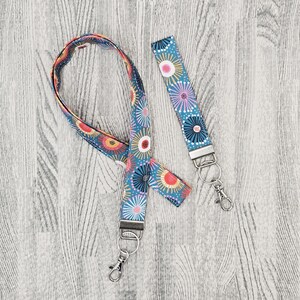 May include: Two matching lanyards with a floral pattern. The lanyards are teal with a repeating design of colourful flowers in red, yellow, pink, and blue. Each lanyard has a silver-coloured metal clasp and key ring.