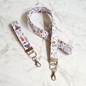 May include: Two white fabric lanyards with a Harry Potter theme. The lanyards feature a white background with illustrations of glasses, wands, letters, and a hat. The lanyards have silver key rings attached.