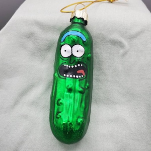 May include: A green glass Christmas ornament shaped like a pickle with a cartoon face. The pickle has a wide, toothy grin, large eyes, and a blue eyebrow.