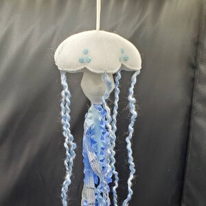 May include: A handmade, whimsical jellyfish ornament. The jellyfish features a white, felt-like cap with blue bead accents and long, flowing blue and white yarn tentacles. A decorative piece for nautical or ocean-themed decor.