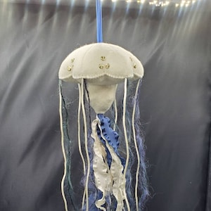 May include: A white felt jellyfish with blue and white fabric tentacles and gold accents. The jellyfish is hanging from a string.
