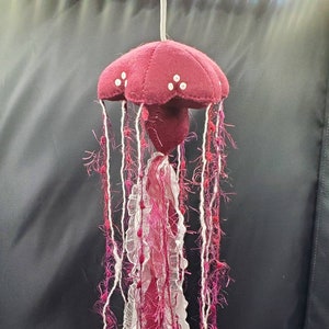 May include: A handmade felt jellyfish mobile with a burgundy bell and white accents. The tentacles are made of white and burgundy yarn and fabric.