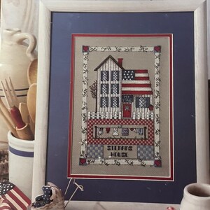 May include: Cross stitch pattern leaflet for a house with a red door and an American flag. The leaflet is titled "Liberty House" by Peggi White. The pattern is for a cross stitch project called "Stars House".