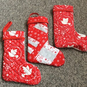 May include: Three red and white Christmas stockings with a white dove design. The stockings are quilted and have a snowflake pattern.