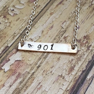 May include: A silver chain necklace with a silver bar pendant that says "♥ 901" on a wood background.