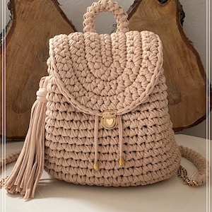 May include: A light pink crocheted backpack with a gold heart-shaped clasp and a long tassel. The backpack has a top handle and a shoulder strap.