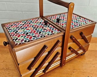 May include: A wooden sewing box with a handle and a multi-coloured floral patterned lid. The box has a tiered design with angled wooden supports and a dark brown handle. The box is on a wooden surface.