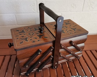 May include: A wooden sewing box with a dark brown finish and floral inlay on the lid. The box has a handle and a multi-tiered design for storage. The box is sitting on a wooden surface.