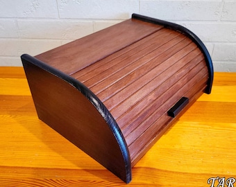 May include: A brown wooden bread bin with a curved, slatted roll-top lid and black trim. The bread bin has a black handle and is set on a wooden surface.