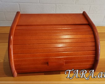 May include: A wooden bread bin with a roll-top lid. The bin is painted a bright orange colour. The lid has a series of horizontal slats. The bin is sitting on a wooden surface. The text "TARA.art" is visible in the lower right corner of the image.