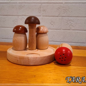 May include: Wooden mushroom salt and pepper shakers with a red mushroom shaker on a wooden base. The shakers have a brown top and a light wood body.