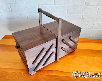 May include: A dark brown wooden sewing box with a handle and hinged lid. The box has angled compartments on the sides. It is displayed on a wooden table. The text "TARA.art" is visible in the bottom right corner. The box is likely for storing sewing supplies.
