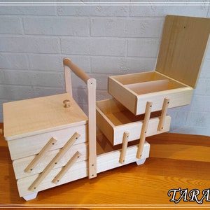 May include: A wooden sewing box with a hinged lid and three drawers. The box has a handle and is made of light wood. The box is open and the drawers are pulled out. The box is sitting on a wooden table.