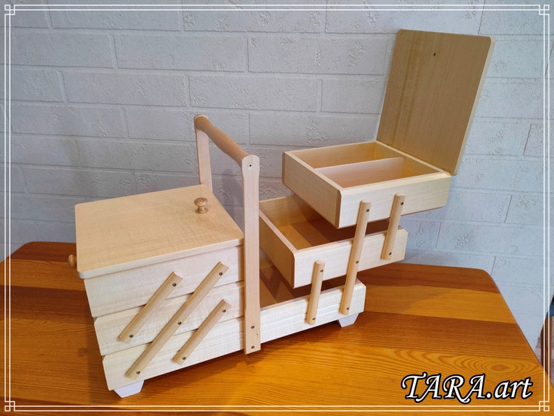 May include: A wooden sewing box with a hinged lid and three drawers. The box has a handle and is made of light wood. The box is open and the drawers are pulled out. The box is sitting on a wooden table.